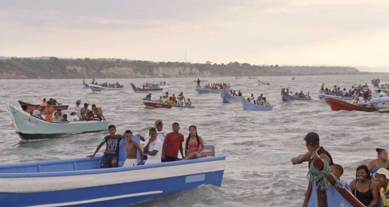 A community of Ecuadorian people are riding in blue boats across the ocean. The ones who are visible in the front are smiling and playfully interacting with each other and the camera.