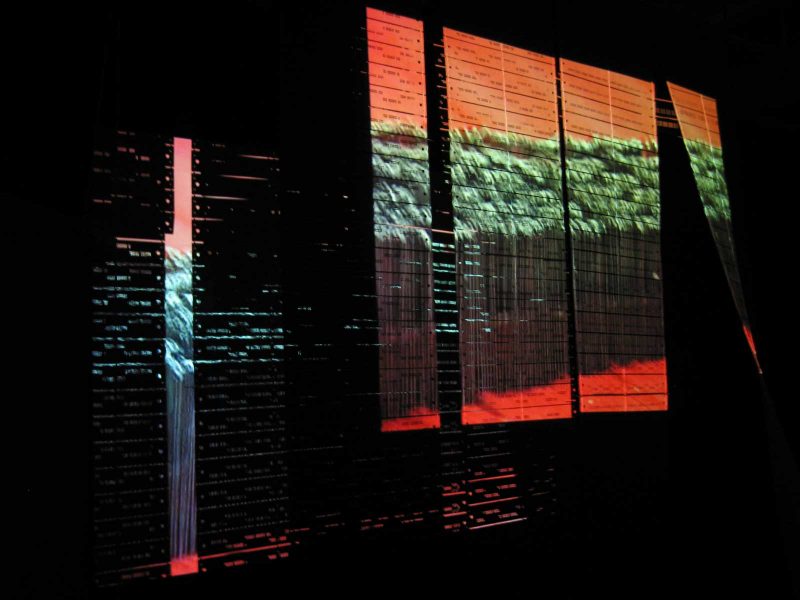 Video installation of a green, organic rectangle on an orange background projected onto paper with patterned holes punched out of it. The light bleeds through the holes in the paper to make an intricate pattern on the wall behind the hung paper.