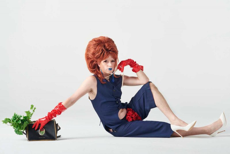 the moon baby in a big red wig, blue jumpsuit, red gloves, and white heels, partially reclined and propping themselves up with one arm.