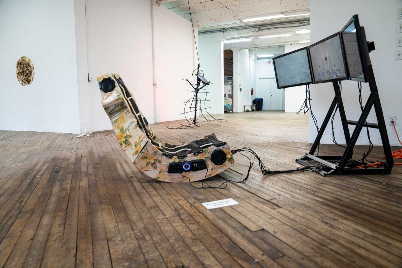 Video installation of a gaming chair, re-upholstered with floral fabric, wired over to, and facing, a black wire frame with three video monitors on it, featuring animated imagery.