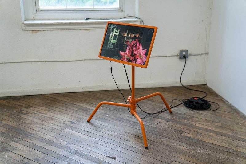 Video monitor showing a dark animated exterior space, with a bouquet of pink lillies in the foreground, installed on an orange metal stand with bent legs.