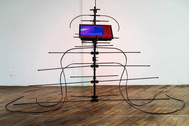 Metal sculpture resembling an abstracted and enlarged TV antenna with a video monitor installed near the top of the antenna. The video shows a field of red, blue, and purple.