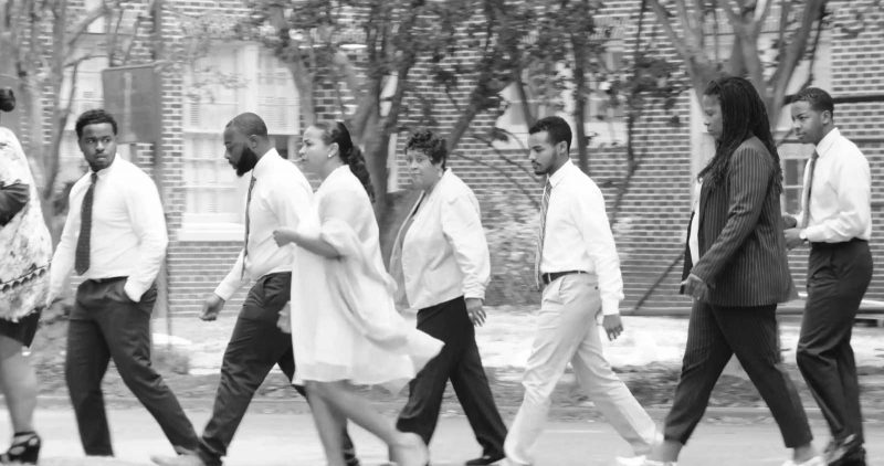 A group of Black women and men wearing formal clothing, chatting as they cross a street.