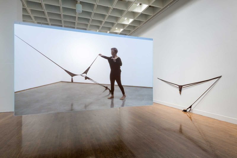 Senga Nengudi video installation showing a woman activating a pantyhose installation. The pantyhose are attached to the wall and floor and the woman stretches a portion of the pantyhose to create another thin line.