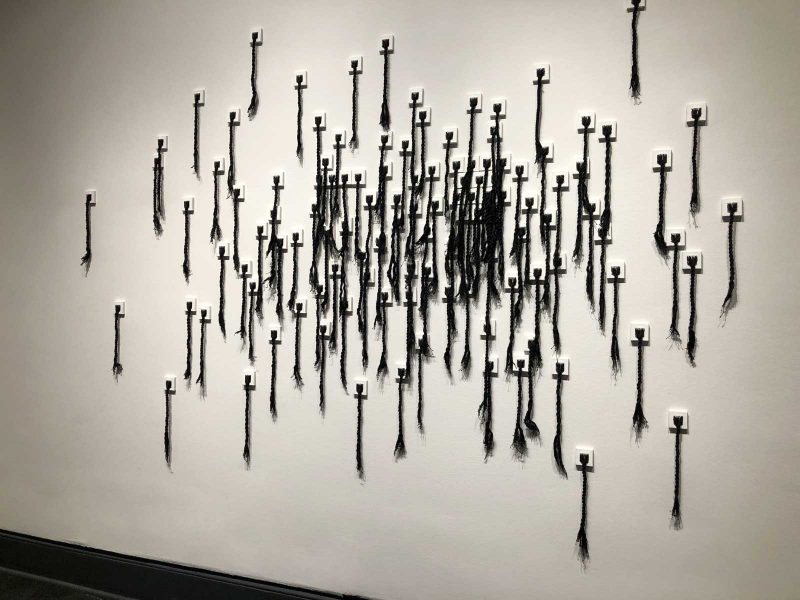 Large wall installation of black fabric braided to resemble Black woman hair that is hung in random clusters on the wall.