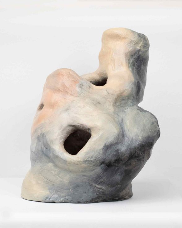 A bulging, slanted, ceramic sculpture with 3 cavernous holes scattered around the hollow mass, which has been glazed with gray, light green, and pink tones, that accentuate the contours of the object.