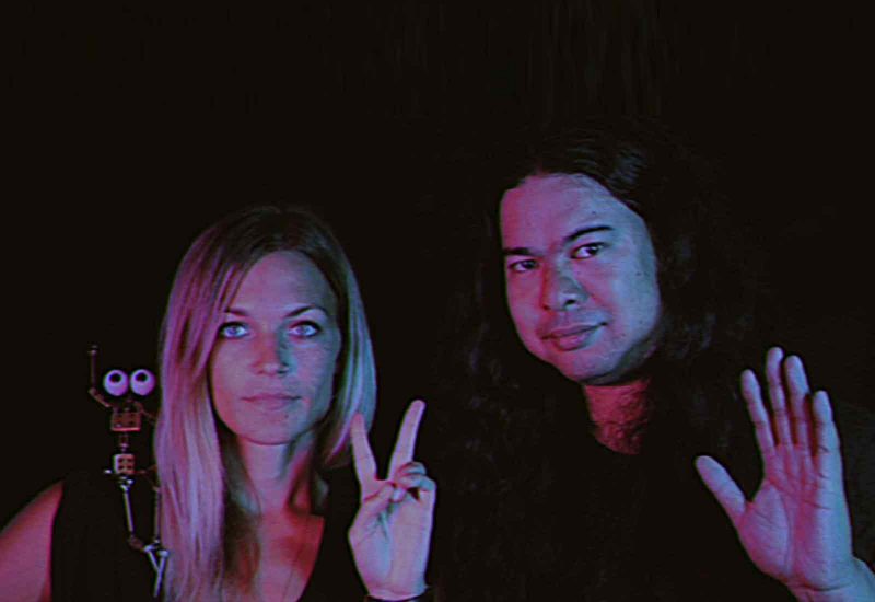 Tamara Hahn, a white woman with blonde hair, with a metal sculpture on her right shoulder, making a peace sign with her left hand; Raymo Ventura, a Filipino-American man with black curly hair, smiling slightly and holding his left hand up with an open palm. 