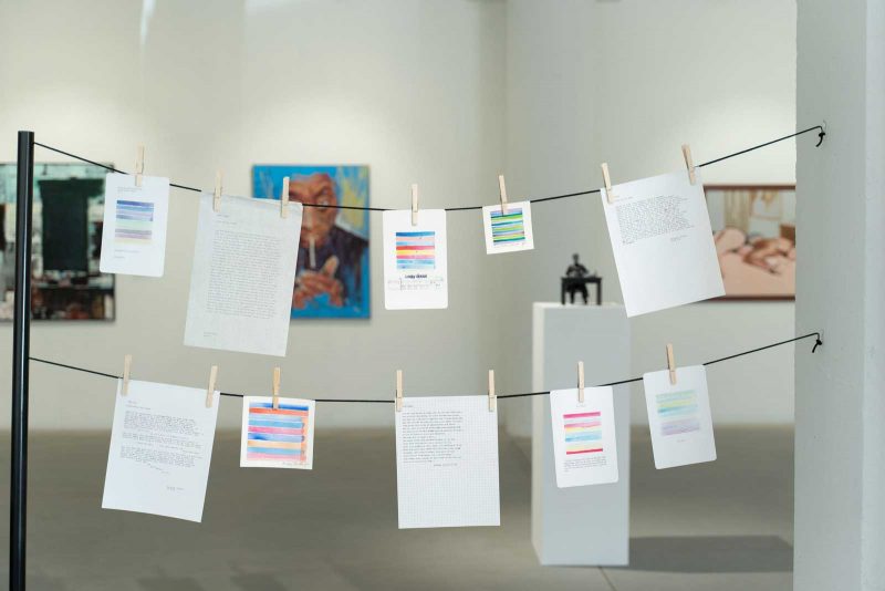 Art Installation of papers filled with writing and color palettes, clipped to black strings, hanging between a black metal bar and a white support beam.