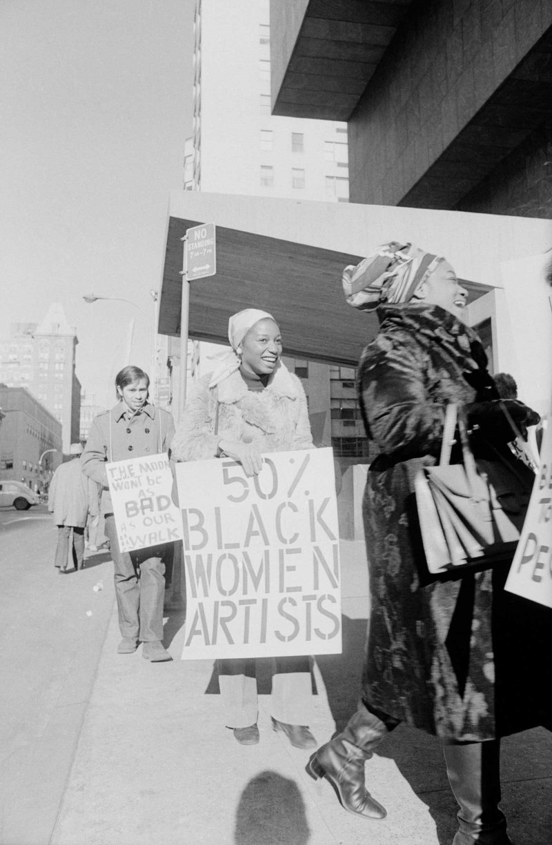 Faith Ringgold (front) and Michele Wallace (back) smiling while they walk in protest; Michelle Wallace holds a sign that reads "50% BLACK WOMEN ARTISTS."