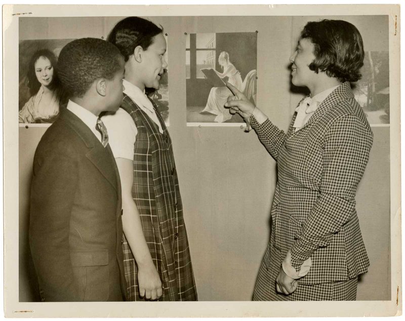 Two students observe as Alma Thomas points to a print of a painting on the wall.