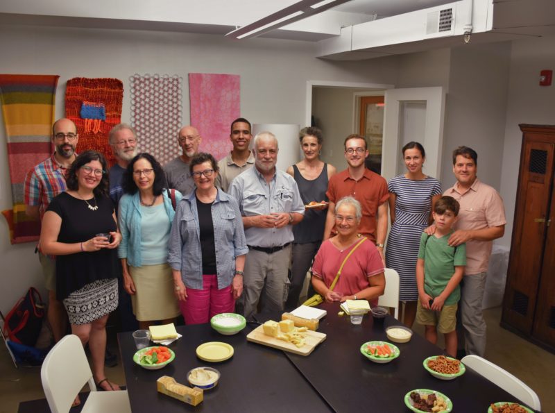 Group of people sitting and standing around a table with food on it, smiling for the camera.