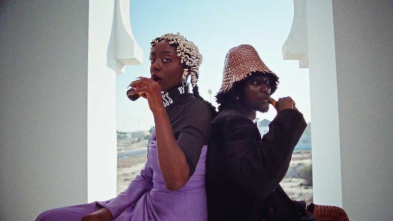 Two young adults, a Black man and a Black woman, sitting back to back and drinking beer in front of an archway with a view of a landscape.