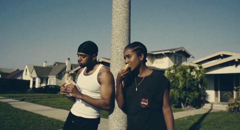 Two friends, a Black man and a Black woman, both wearing tank taps, lean against a concrete pole on a suburban street as they eat food together.