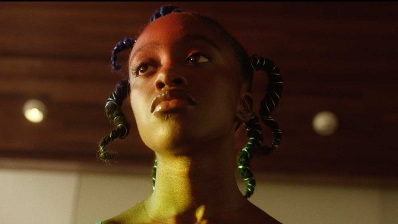 Close-up of a young Black woman with thick braids wrapped silver wire, who is lit with red light near the top of her face and yellow-is green light on her chin and neck. 