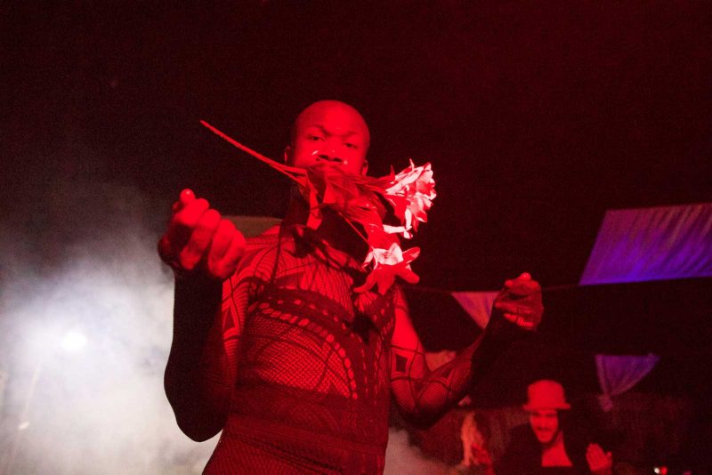 Arien Wilkerson, a Black man with a shaved head, staring into the camera and holding a bouquet of flowers in his mouth in a dark, foggy room with red lighting.