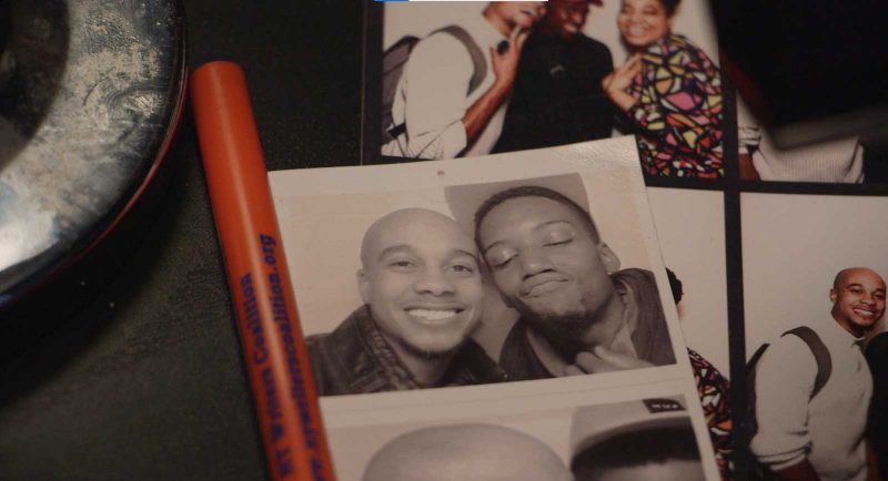 Video still, showing the corner of an ashtray, an 'NY Writers Coalition' pen, and two sets of photos, in which one Black man appears in both. In one, he is posing and smiling with another Black man in a photo booth. In the other, he is posing with another Black man and a Black woman, and they are smiling and making silly faces.