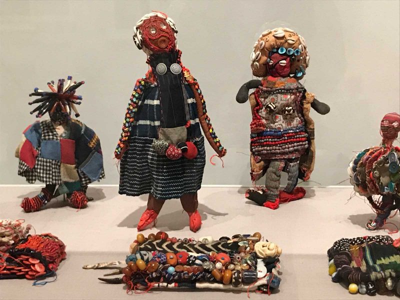 7 sculptures-- three of books, three of figures, all of which are constructed out of fabric, beads, and buttons-- arranged in two lines (books in front; figures in back; in a museum display.