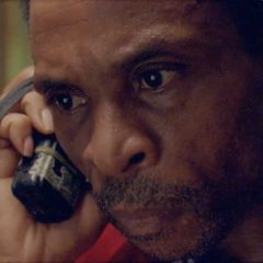 Close-up on a middle-aged Black man with short salt-and-pepper hair and facial hair, holding a walkie talkie up to his ear and looking concerned.