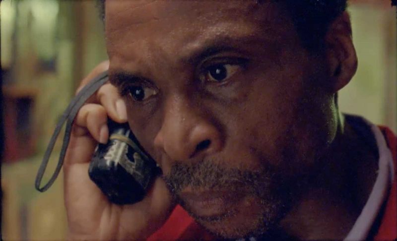 Close-up on a middle-aged Black man with short salt-and-pepper hair and facial hair, holding a walkie talkie up to his ear and looking concerned.