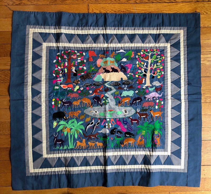 Blue fabric artwork embroidered with many animals surrounding a stream, some drinking and others resting on a nearby hill or mountain.