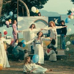 Five women in white dresses perform a musical number in front of a rainbow banner, surrounded by colorful balloons and groups of friends socializing nearby.