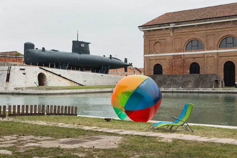 A big colored beach ball sits near a canal, with two beach chairs to the right, a boat building in the rear and a black submarine sitting out of the water.