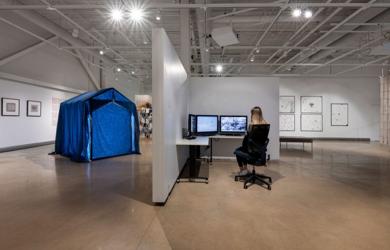 Art gallery with multiple works installed inside of it, including a large glittery blue tent, and a L-shaped desk with three computer monitors, playing videos; a person in a swivel chair sits and watches them.