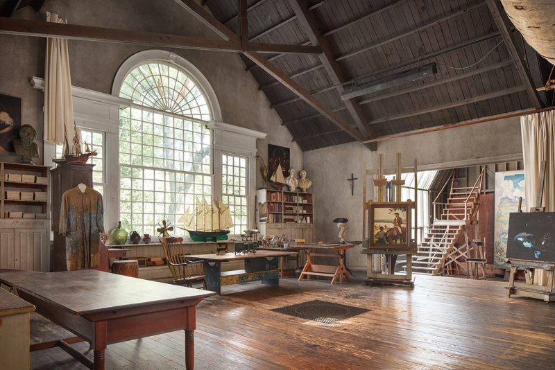 Large room with huge white windows, high ceilings with exposed wooden beams and supports, and hardwood floors, with multiple painting easels, work benches, and sculptures throughout the room. 
