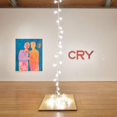 Gallery space with an installation of hanging lightbulbs in the center, which descend in two lines into a bundle that rests on a podium; with two hanging works on the wall behind: an abstract painting of figures in a side embrace (left) and a hanging sculpture of stylized red letters that spell "CRY" (right).