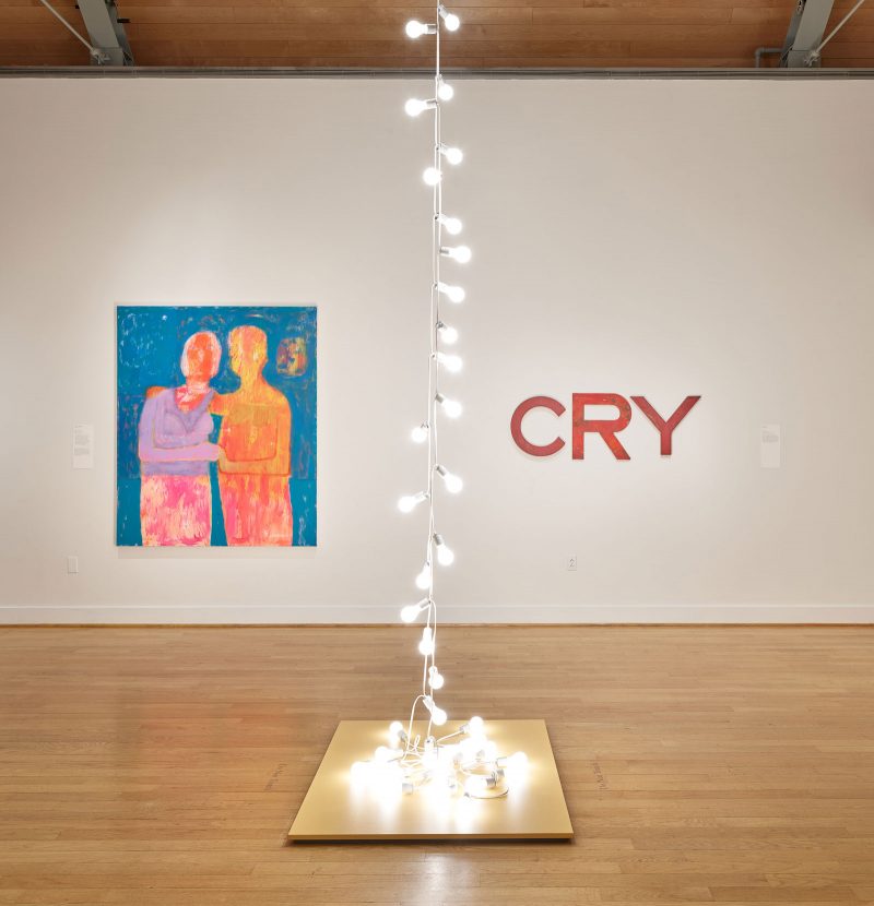 Gallery space with an installation of hanging lightbulbs in the center, which descend in two lines into a bundle that rests on a podium; with two hanging works on the wall behind: an abstract painting of figures in a side embrace (left) and a hanging sculpture of stylized red letters that spell "CRY" (right).