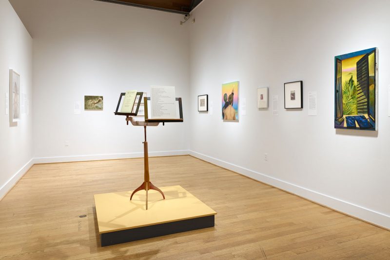 A podium in the center of a gallery space raises a single music stand base, from which there are two stand attachments, tilted away from each other at a 45 degree angle, each with paper and written text sitting on it; behind, many 2D artworks hang on the walls.