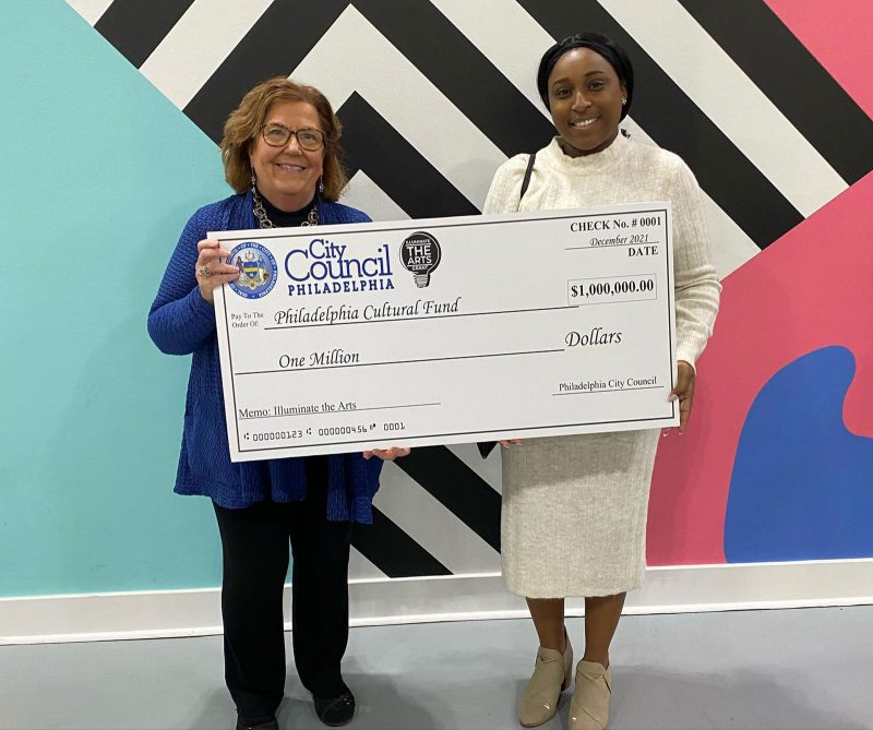 Two women hold up a poster of a check for $1 million, standing in front of a colorful wall.