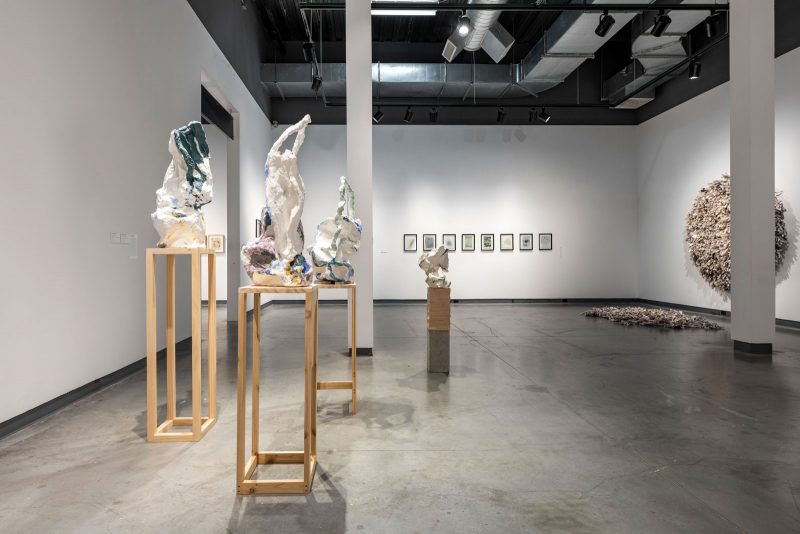 Installation view of a museum exhibition, with abstract sculptures on wooden pedestals in the foreground, and other hanging works installed on the walls behind.