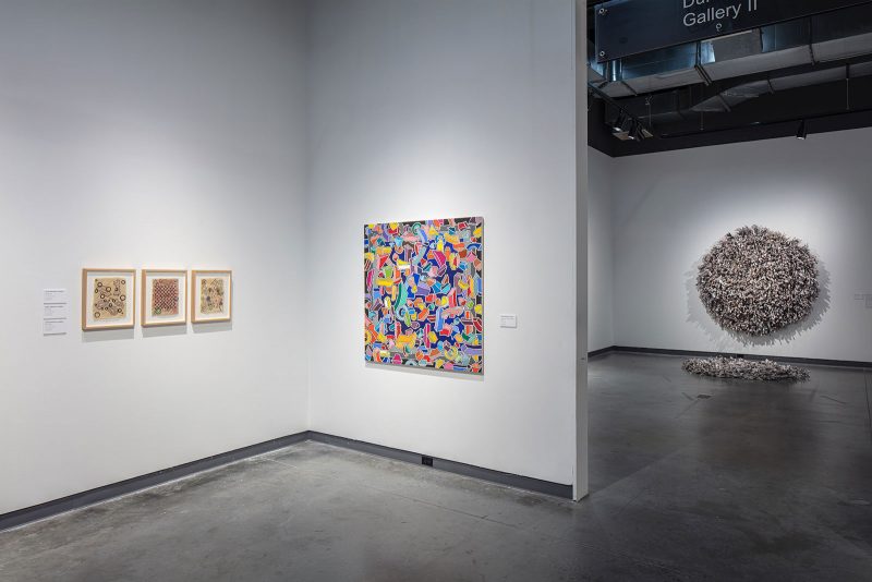 Installation view of a museum exhibition, with a front section featuring three framed small earth-toned works on paper, and a larger, busy, geometric and colorful, abstract painting; in the farther section of the gallery, a large textured circular hanging wall piece dominates the wall, with its material spilling onto the floor in front of it.