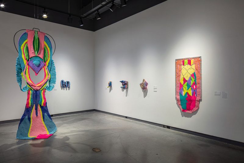 Installation view of a museum exhibition, with a large vaginal colorful fabric piece in the foreground, and smaller dimensional fabric works hanging on the walls nearby, plus one medium-sized two dimensional hanging colorful fabric piece. 