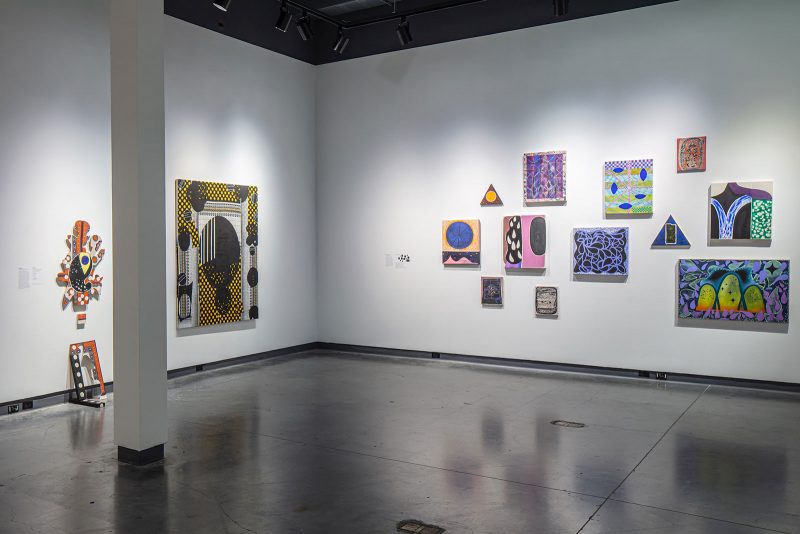 Installation view of a museum exhibition, featuring abstract paintings on two walls; on the left, a uniquely shaped work that spills onto the floor and the largest painting, which is black and yellow; on the right, a cluster of smaller works, with similar cool color palettes, amassed into the center of the wall.