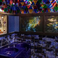 A photo of a bathroom shows two sinks colored purple, in a room whose walls are tiled in shades of purple, and with an altar-like installation on the left wall and two images of the aurora borealis on the right wall.