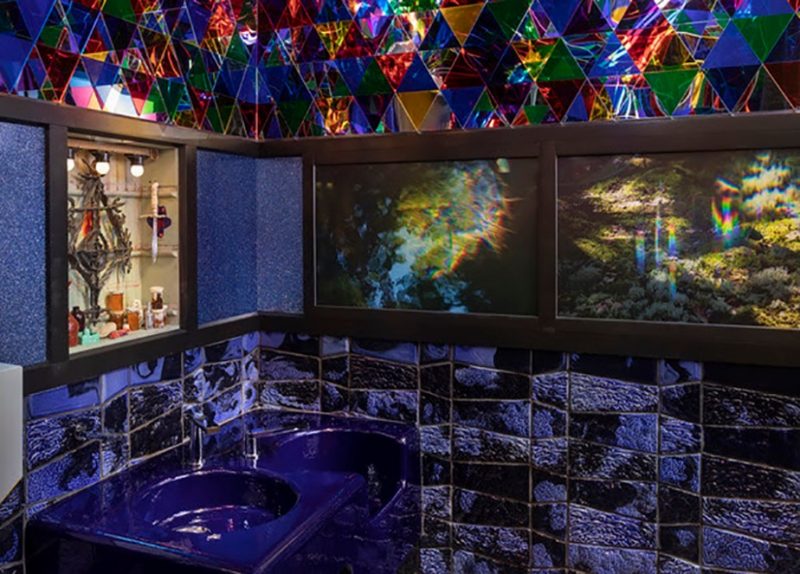 A photo of a bathroom shows two sinks colored purple, in a room whose walls are tiled in shades of purple, and with an altar-like installation on the left wall and two images of the aurora borealis on the right wall.