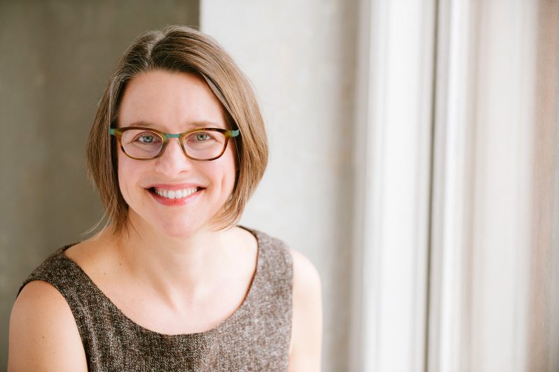 Lynn Tomaszewski, a white woman with chin-length light brown hair, wearing green and tortoise shell-framed glasses, and a brown, textured, sleeveless blouse.