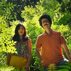 A man (right) and woman (left) standing in a lush woods area, both smiling slightly and looking into the camera.