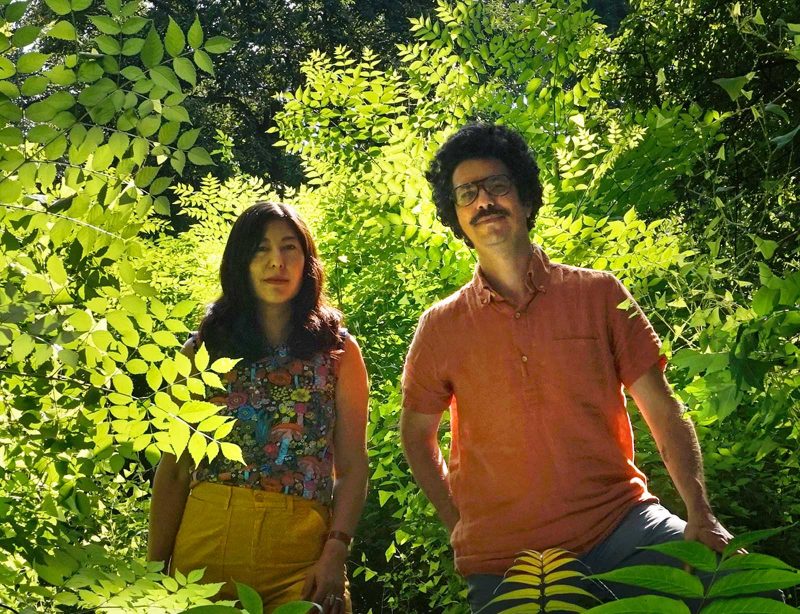A man (right) and woman (left) standing in a lush woods area, both smiling slightly and looking into the camera. 