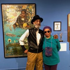 An African American man and woman stand together in a friendly pose in front of a large colorful painting on a dark blue wall in a museum