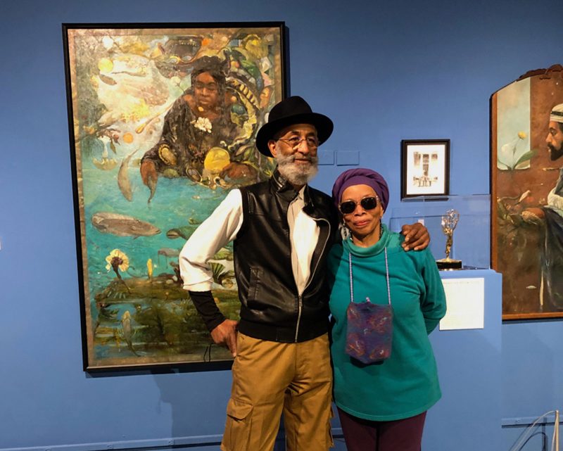 An African American man and woman stand together in a friendly pose in front of a large colorful painting on a dark blue wall in a museum