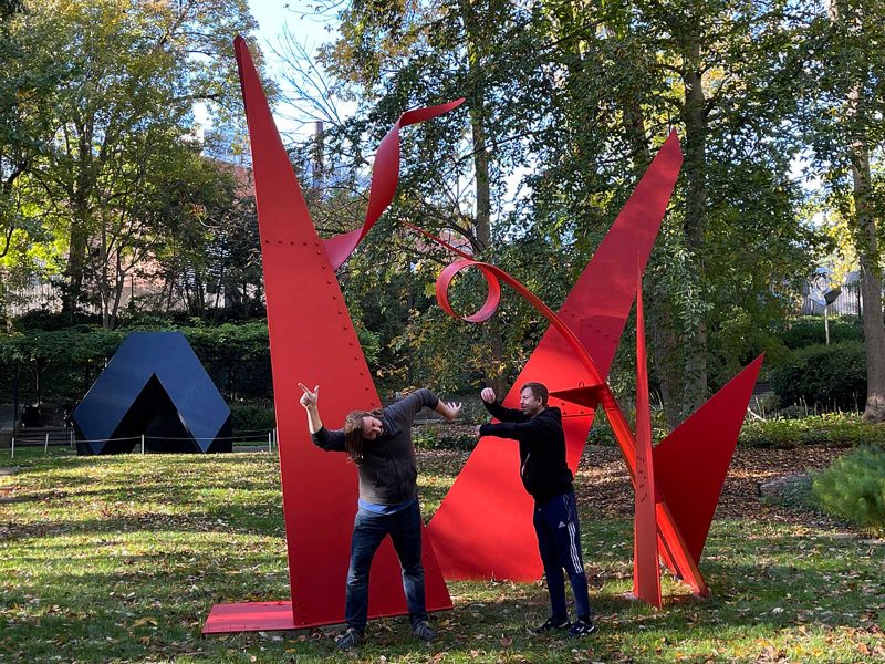 Two men posting with their limbs in shapes that resemble a large, red, abstract outdoor sculpture behind them.