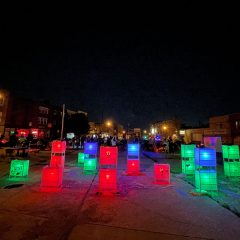 Glowing boxes and vertical rectangles are installed in an empty lot, where people have convened behind it, near a projector screen and a band.