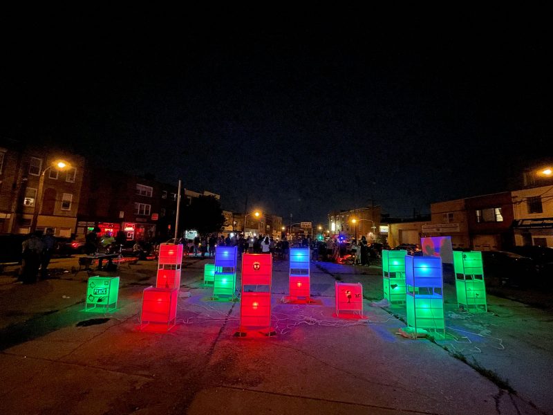 Glowing boxes and vertical rectangles are installed in an empty lot, where people have convened behind it, near a projector screen and a band. 
