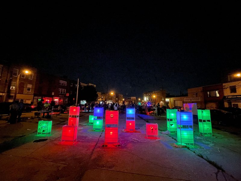 Glowing boxes and vertical rectangles are installed in an empty lot, where people have convened behind it, near a projector screen and a band.