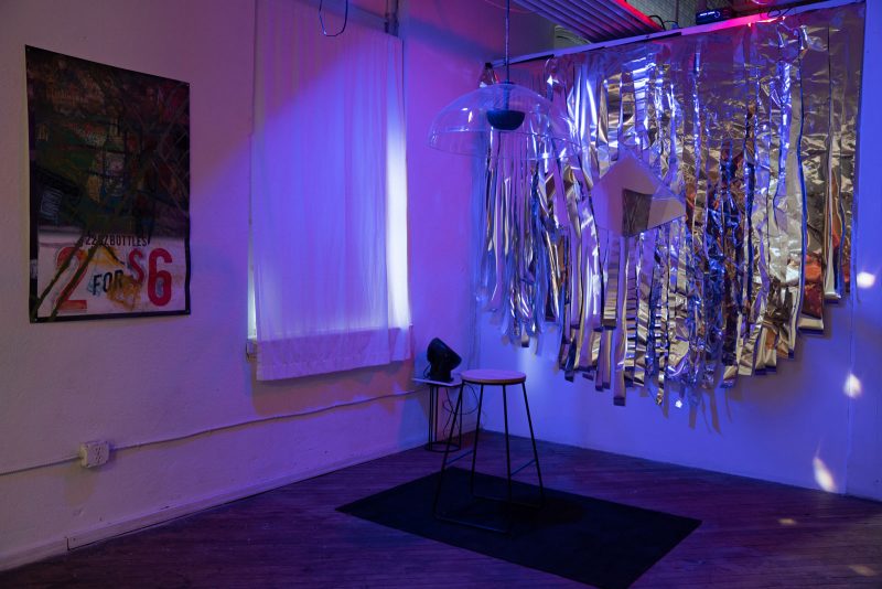 Installation view of a stool on a black carpet, underneath a salon hair-dryer shaped plastic sculpture, with a mirror and large silver sparkly tinsel (which is moving due to a nearby fan) hanging on the wall behind it.