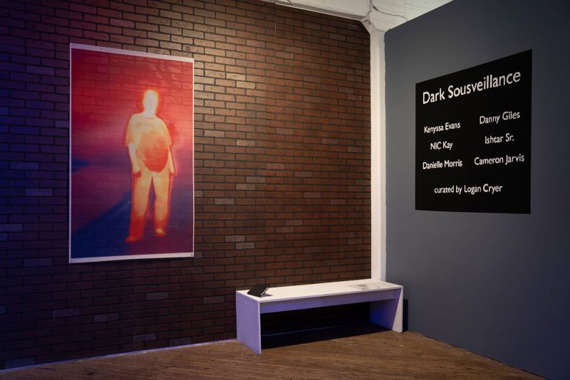 A portrait of a person depicted in thermal heat colors, printed onto a sheer mesh, hangs on a wall that's been covered with faux brick (left); on a neighboring gray wall is a black square with the exhibition title "Dark Sousveillance" and the 6 featured artists' names.