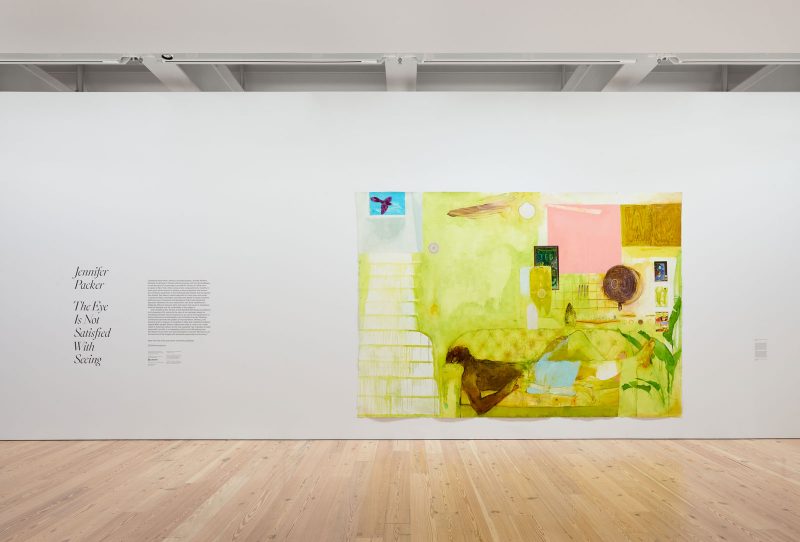 Installation view of the entrance to Jennifer Packer's Whitney exhibit, with wall text and a large yellow painting on paper of a black man lying on a sofa.